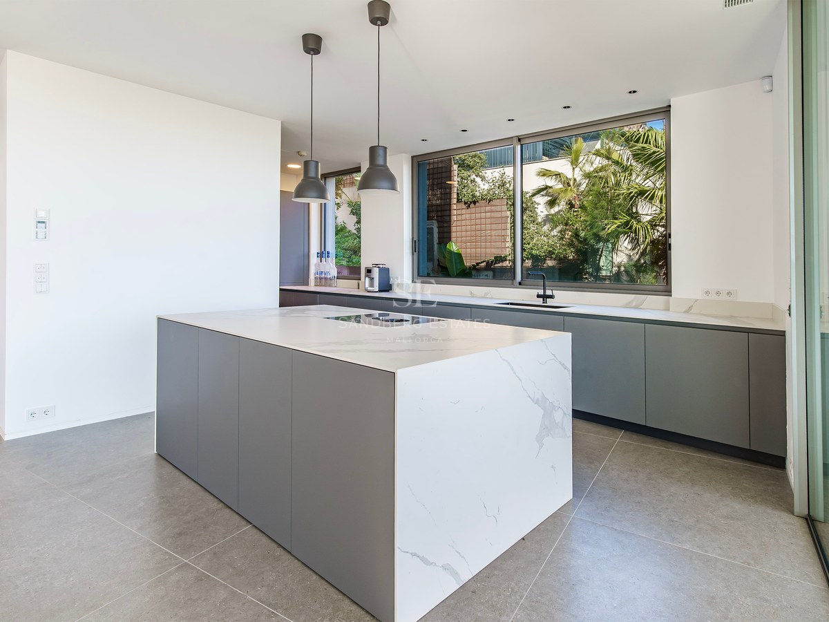 Cocina moderna con una gran isla de mármol blanco, gabinetes grises y ventana con vista a plantas tropicales.