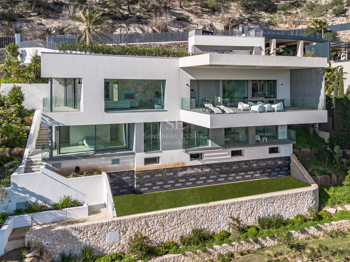 Villa moderna blanca de varias plantas con grandes ventanales y terrazas construida en una ladera rocosa.