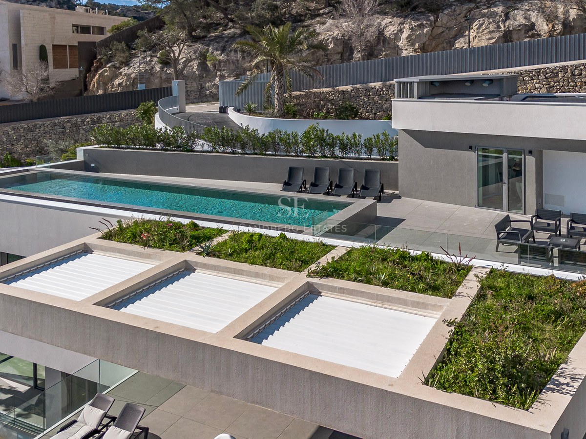 Vista aérea de una villa moderna gris con piscina infinita, tumbonas y vegetación en la azotea frente a una colina.