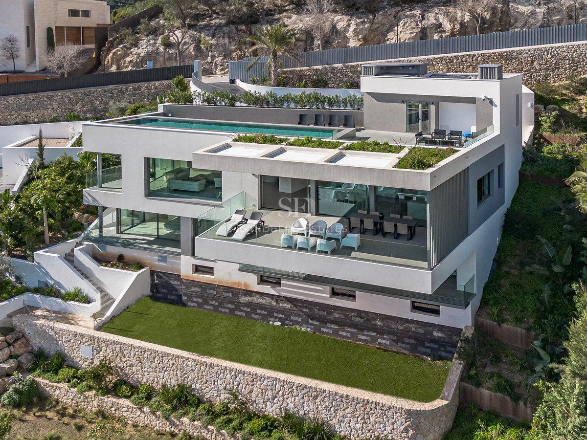 Vista aérea de una villa moderna blanca de varias plantas con piscina infinita en la azotea y terrazas de cristal.