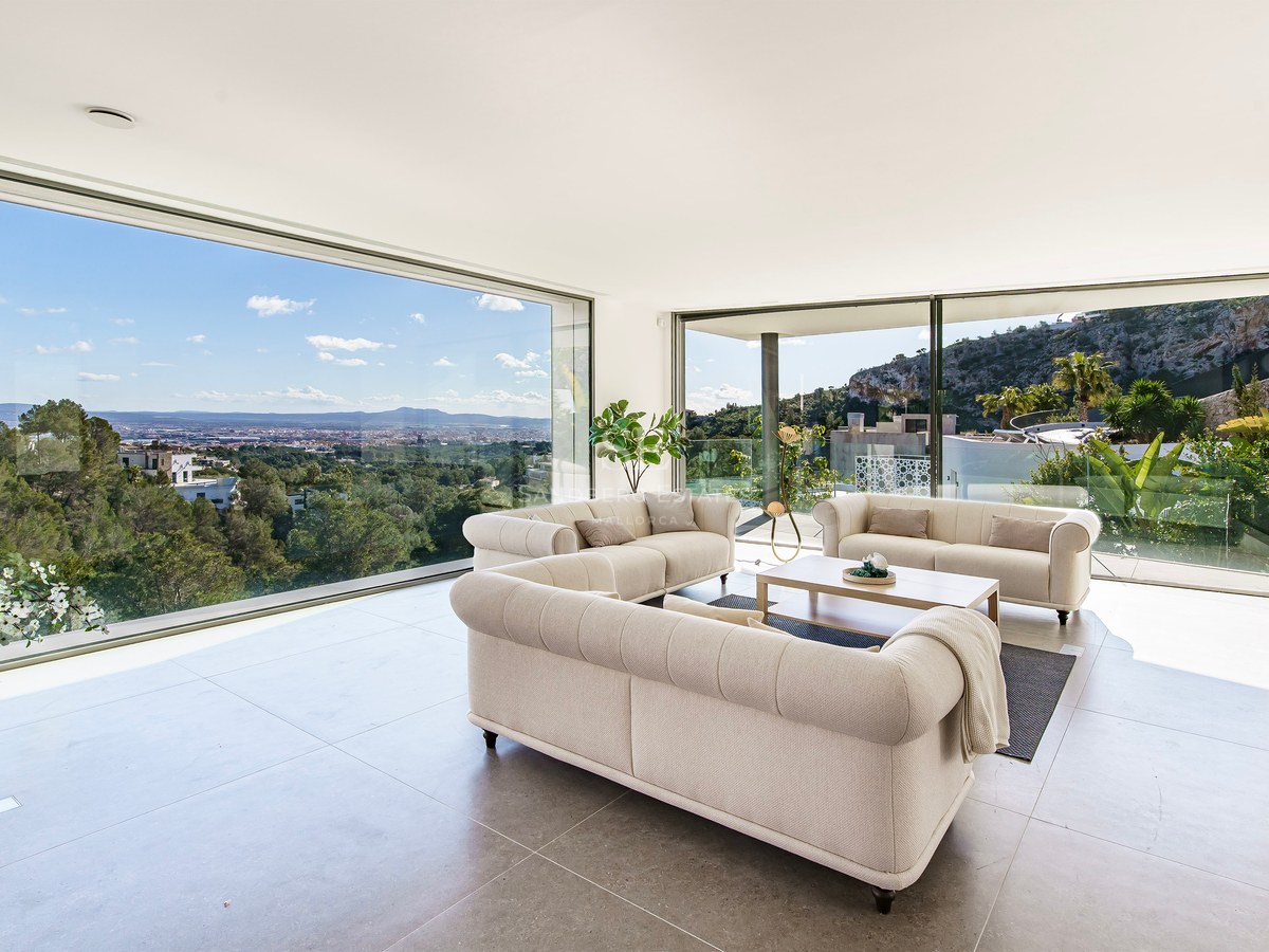 Salón moderno con tres sofás beige y grandes ventanales que muestran vistas a la ciudad y las montañas.