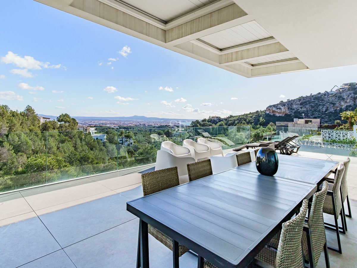 Terraza moderna con mesa de comedor y barandillas de cristal con vistas a un bosque y la ciudad lejana.