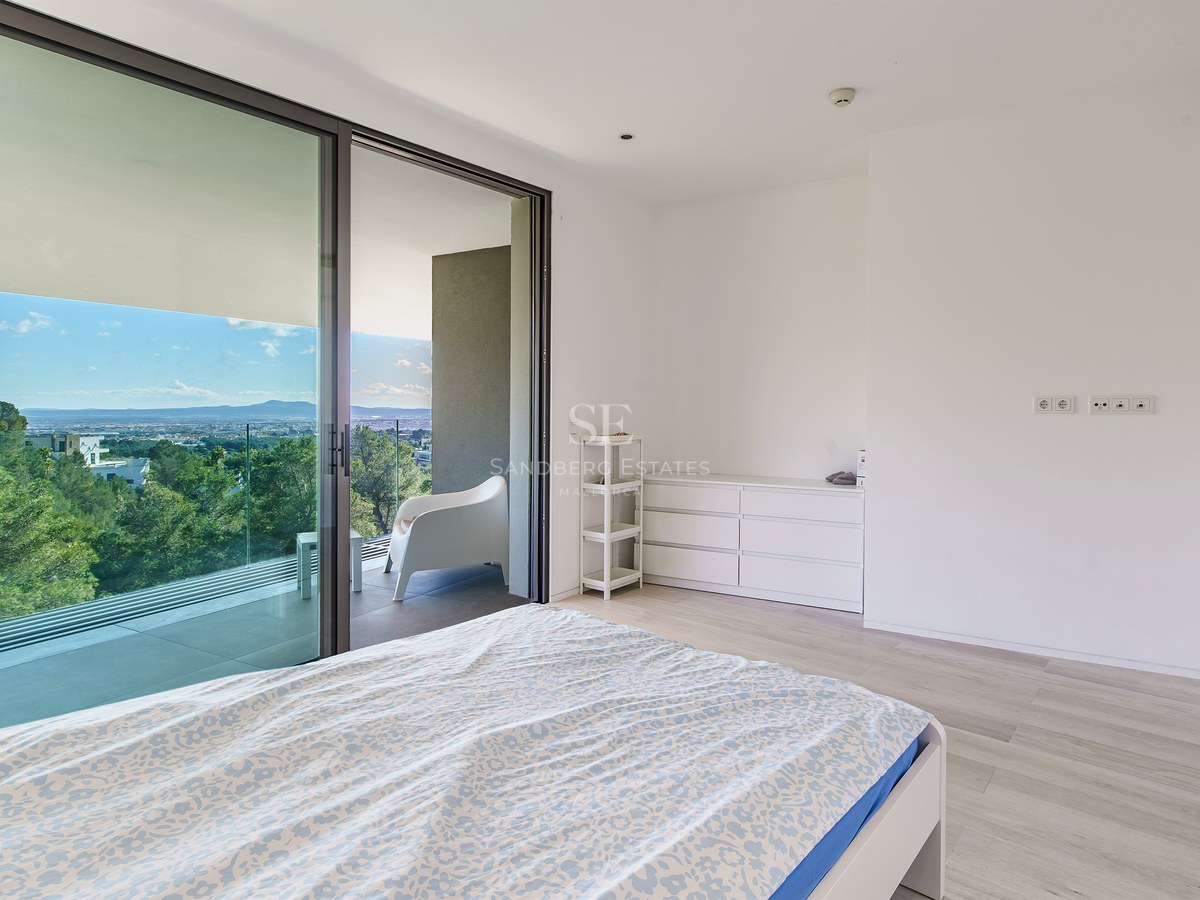 Dormitorio blanco minimalista con grandes puertas de cristal a una terraza con vistas al bosque.