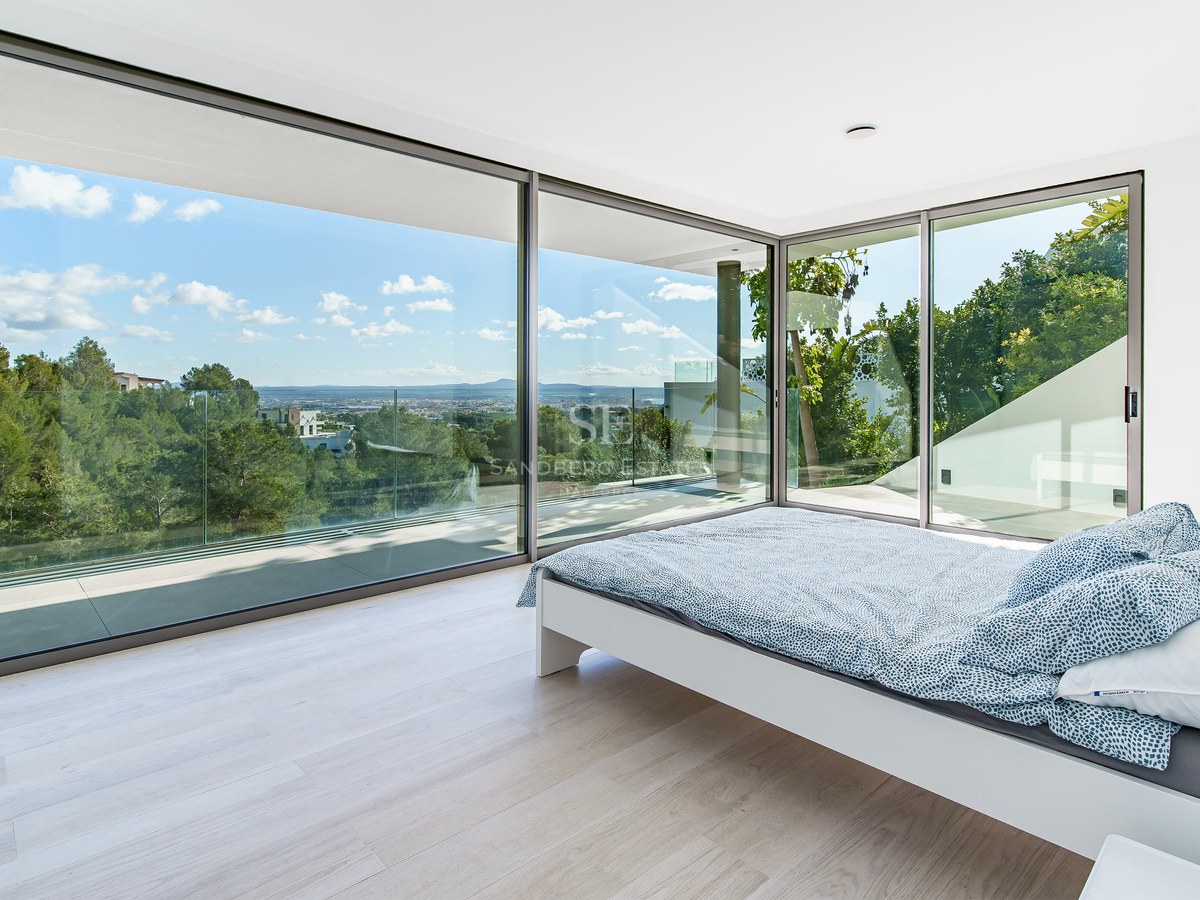 Dormitorio moderno con cama blanca y grandes ventanales con vistas a un bosque verde y la ciudad al fondo.