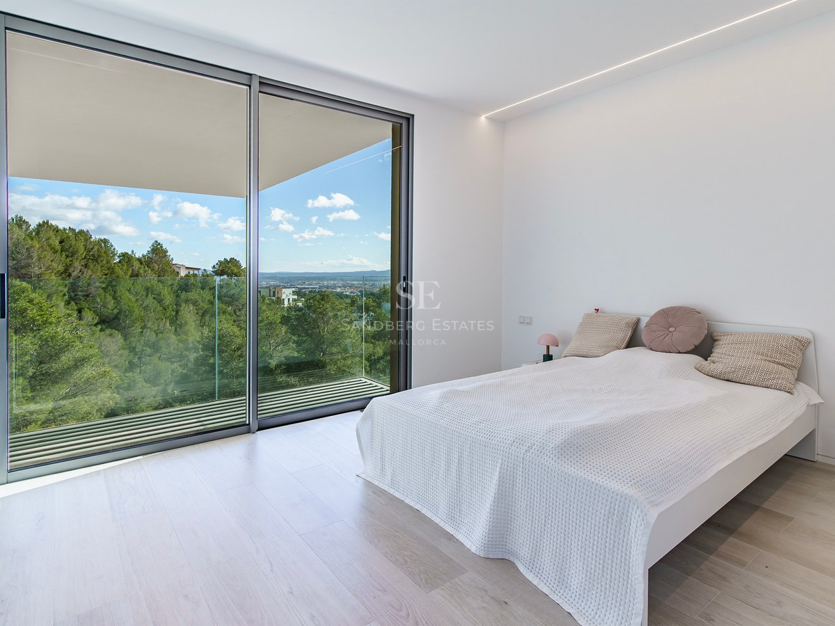 Dormitorio minimalista luminoso con grandes puertas correderas de cristal que dan a un balcón con vistas a colinas verdes.