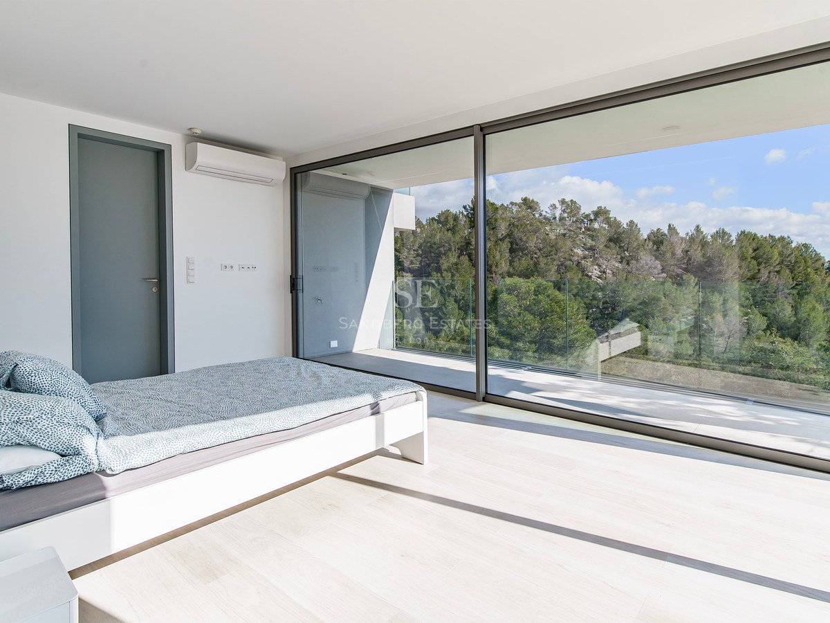 Dormitorio luminoso con ventanales de suelo a techo que dan a un balcón con vistas a un frondoso bosque verde.