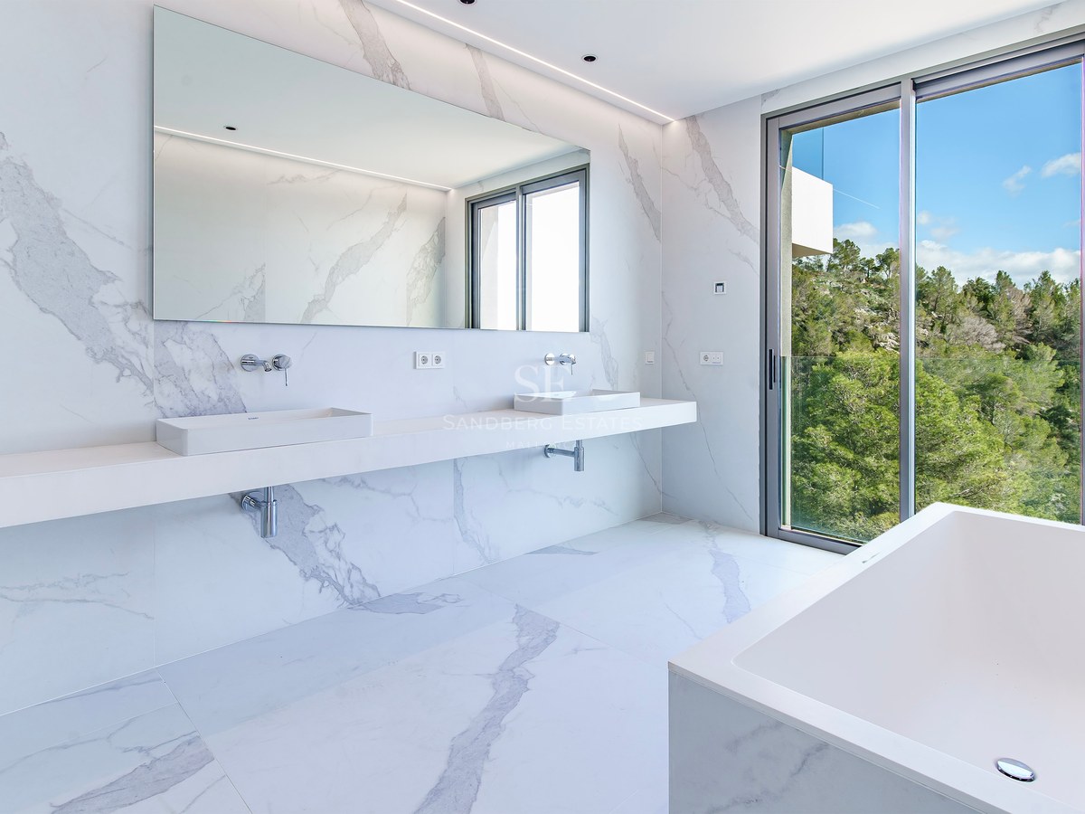 Baño principal luminoso con paredes y suelos de mármol blanco, lavabo doble, bañera y vistas al bosque.