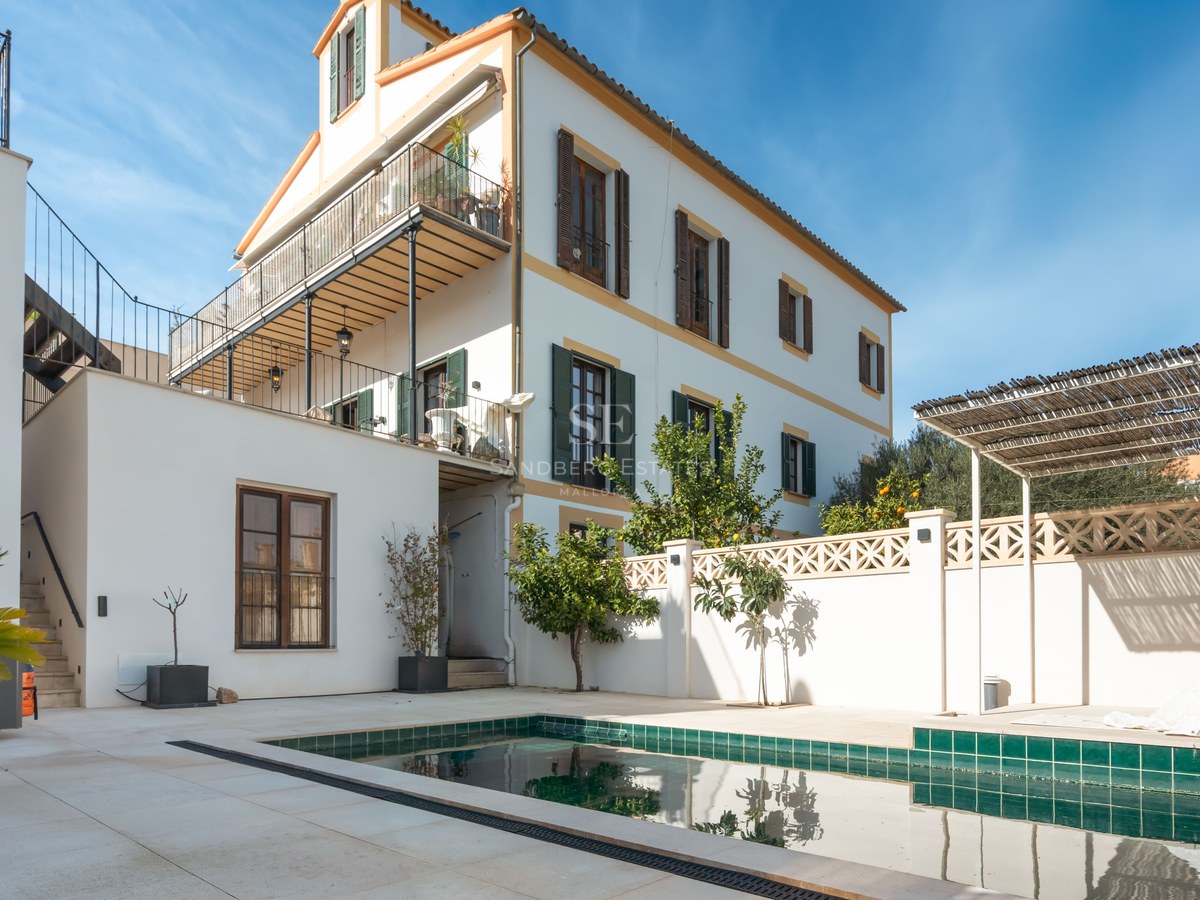 Grönkaklad pool på en vit stenterrass bredvid en traditionell villa med fönsterluckor i trä.