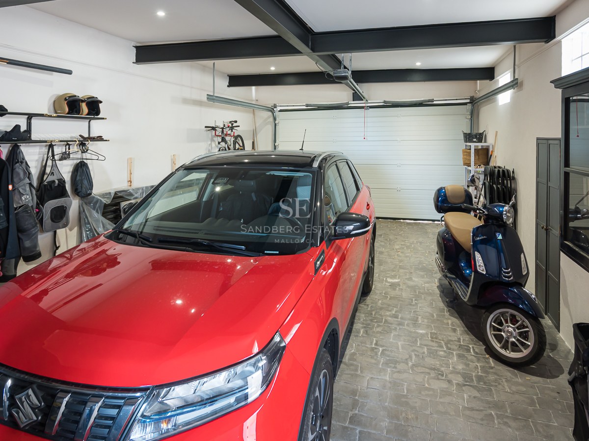 Ljust garage med röd bil, blå skoter, småstensgolv och svarta stålbalkar i taket.