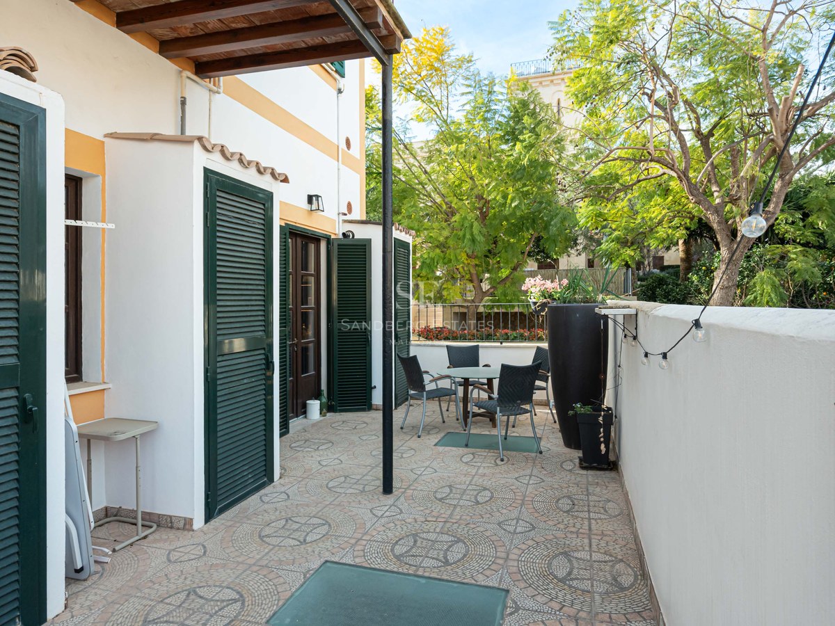 Utomhusterrass med bord, stolar, mönstrat klinkergolv och gröna fönsterluckor under en pergola.