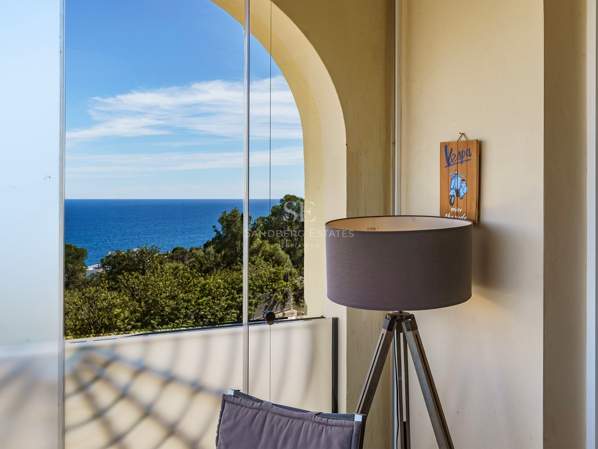 A covered terrace with frameless glass curtains looking out over pine trees to the blue Mediterranean sea.