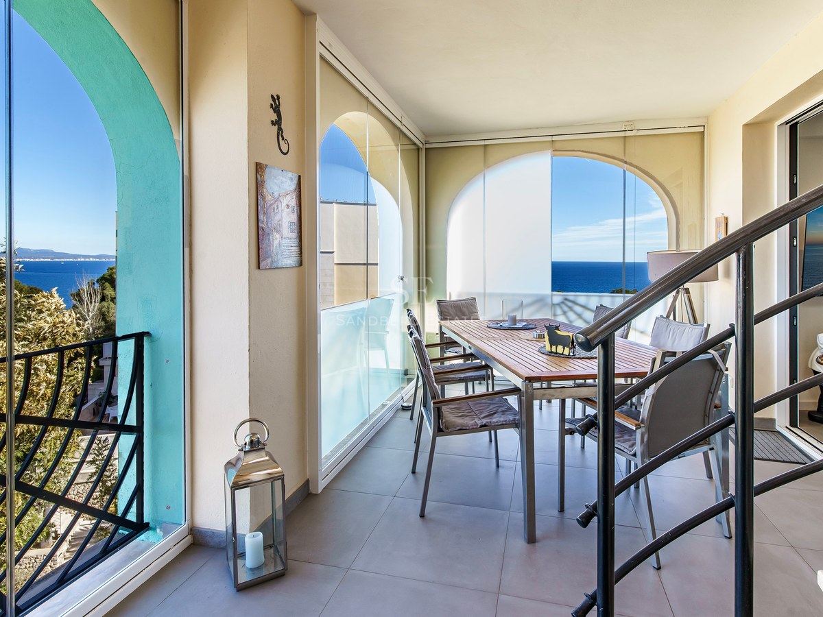 Covered dining terrace with wooden table, glass curtains, and views of the blue Mediterranean sea through arched openings.