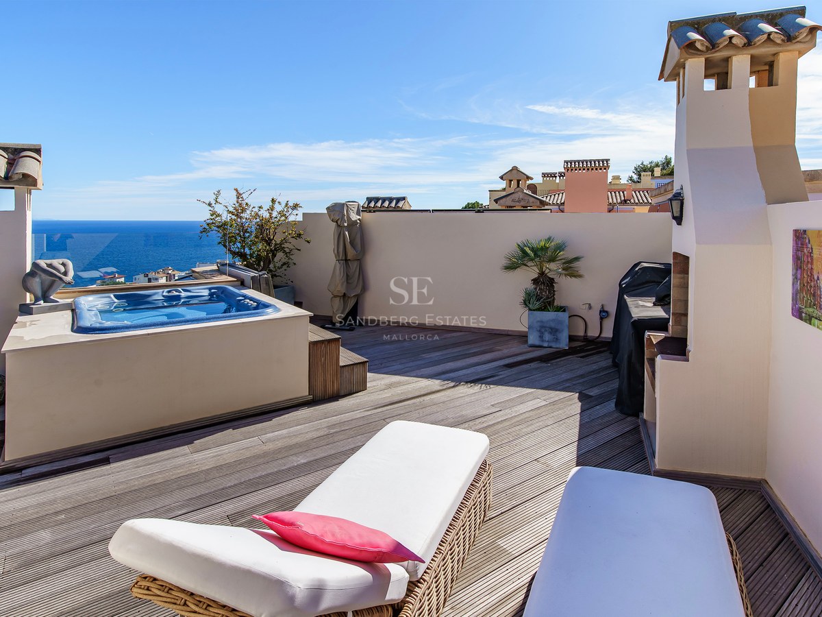 A sunny rooftop terrace with wooden decking, a built-in hot tub, white sun loungers, and panoramic views of the Mediterranean.