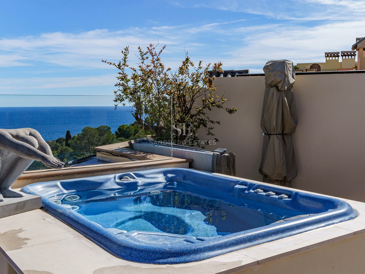 Blue built-in jacuzzi on a stone terrace with glass railings overlooking the Mediterranean Sea and a stone sculpture.