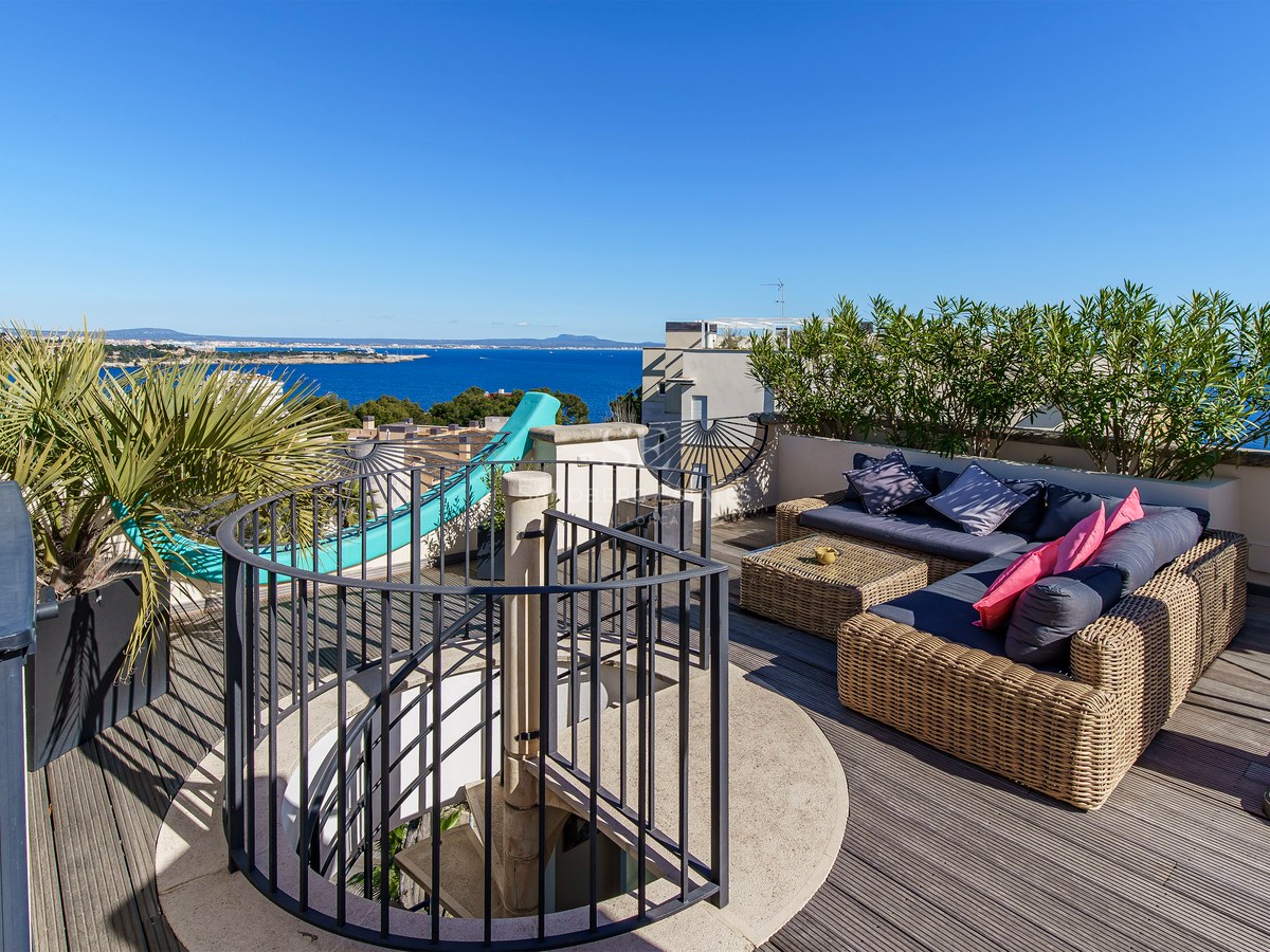A sun-drenched wooden rooftop terrace with wicker sofas and a spiral staircase, overlooking the Mediterranean Sea.