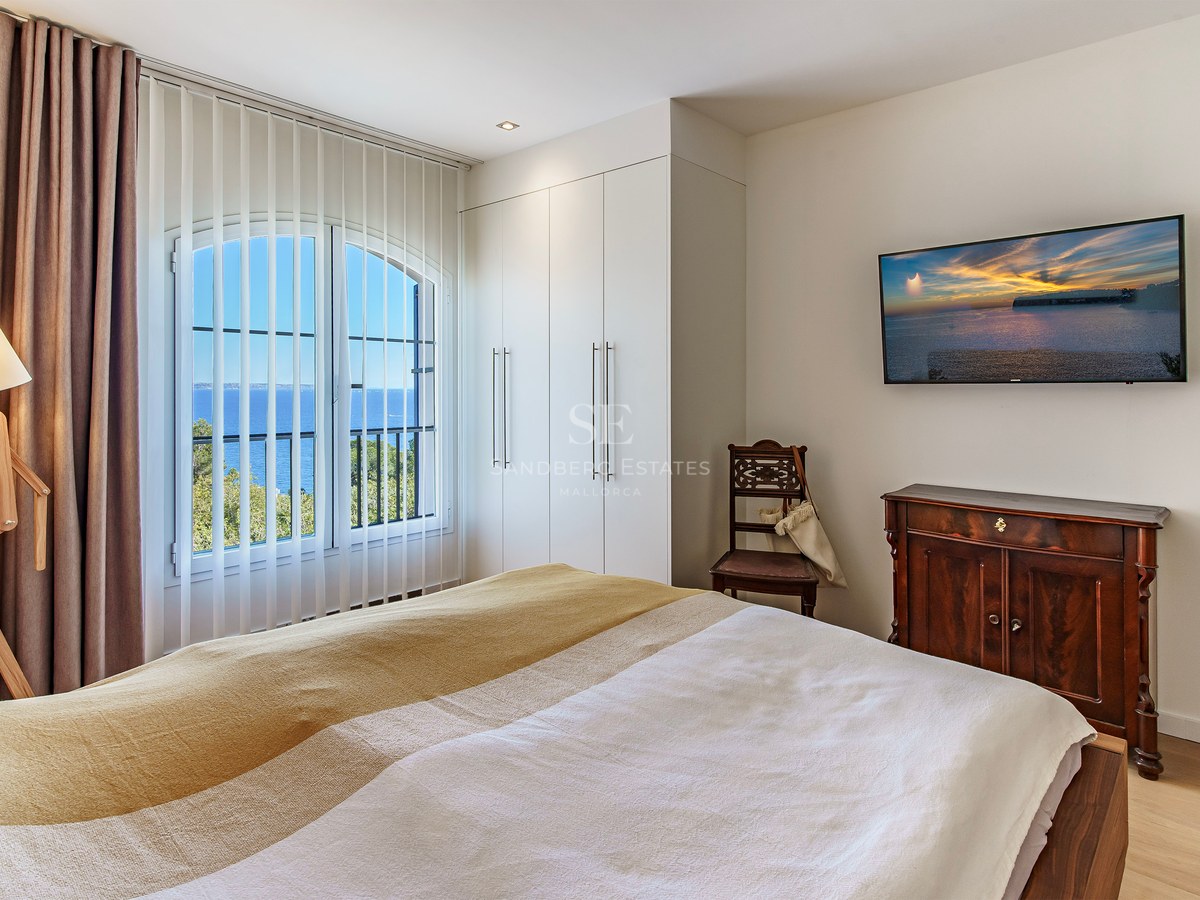 Bright bedroom with an arched window framing a Mediterranean sea view, featuring white wardrobes and wooden flooring.