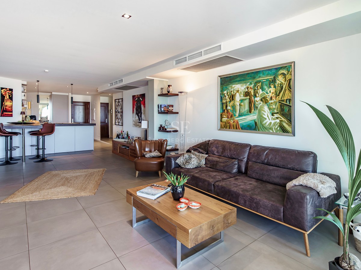 Modern living room featuring a brown leather sofa, wooden coffee table, large-format grey tiles, and integrated kitchen.