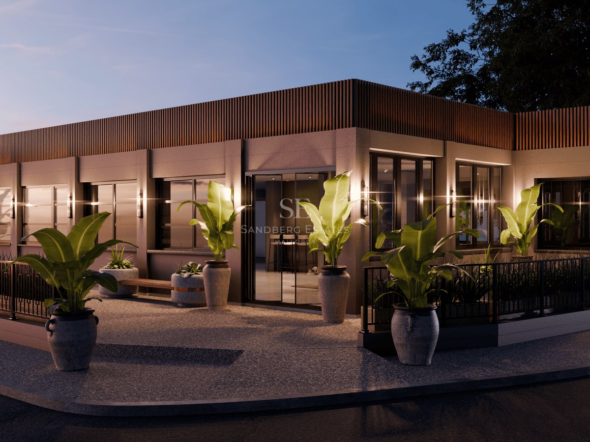 Modern single-story building at dusk with wooden slats, large windows, and tropical potted plants along a stone walkway.