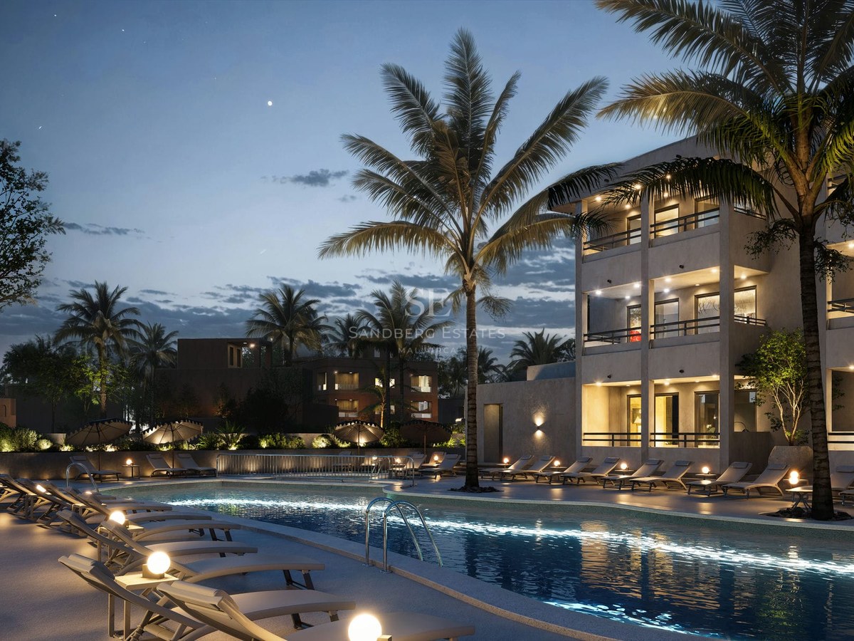 Illuminated swimming pool at night with palm trees, sun loungers, and a modern multi-story building under a starry sky.
