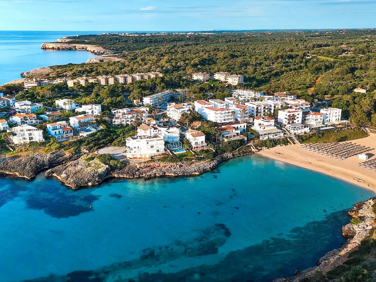 Aerial drone view of a turquoise bay with a sandy beach surrounded by white Mediterranean buildings and lush green forest.