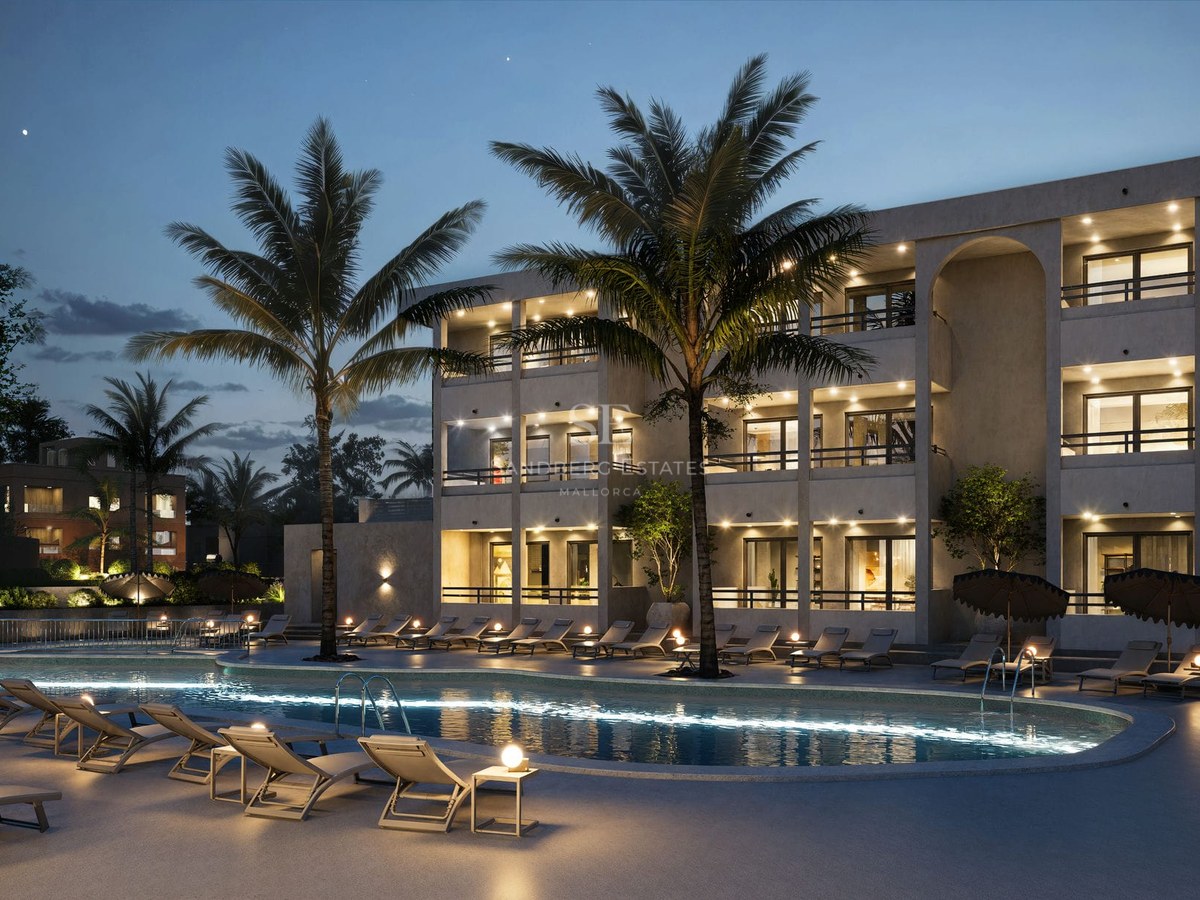 Large illuminated swimming pool with sun loungers and palm trees in front of a modern three-story building at dusk.