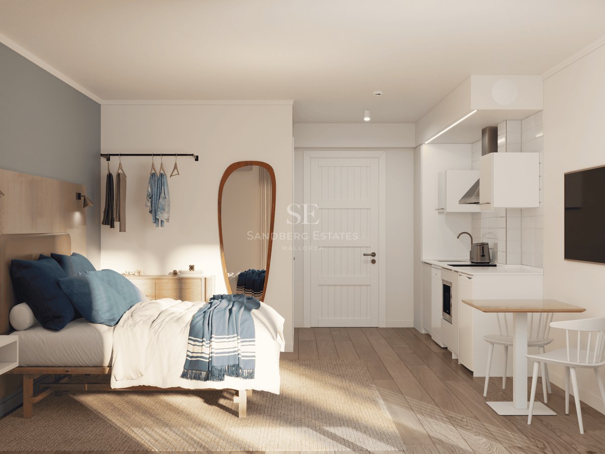 Bright studio apartment featuring a bed with blue accents, a compact white kitchen, and light wood flooring.