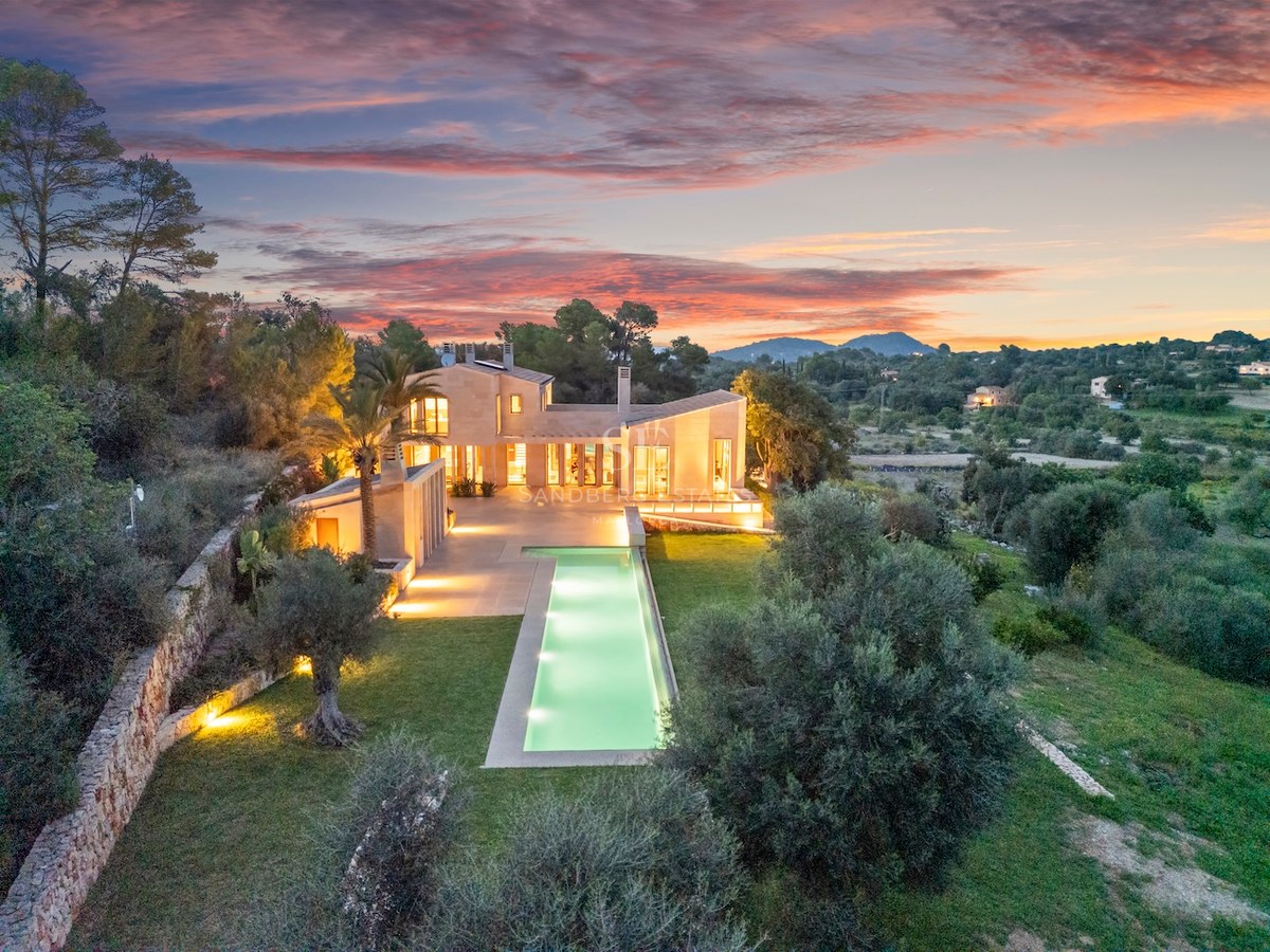 Aerial view of a luxury villa with a long glowing pool at sunset, surrounded by trees and stone walls.