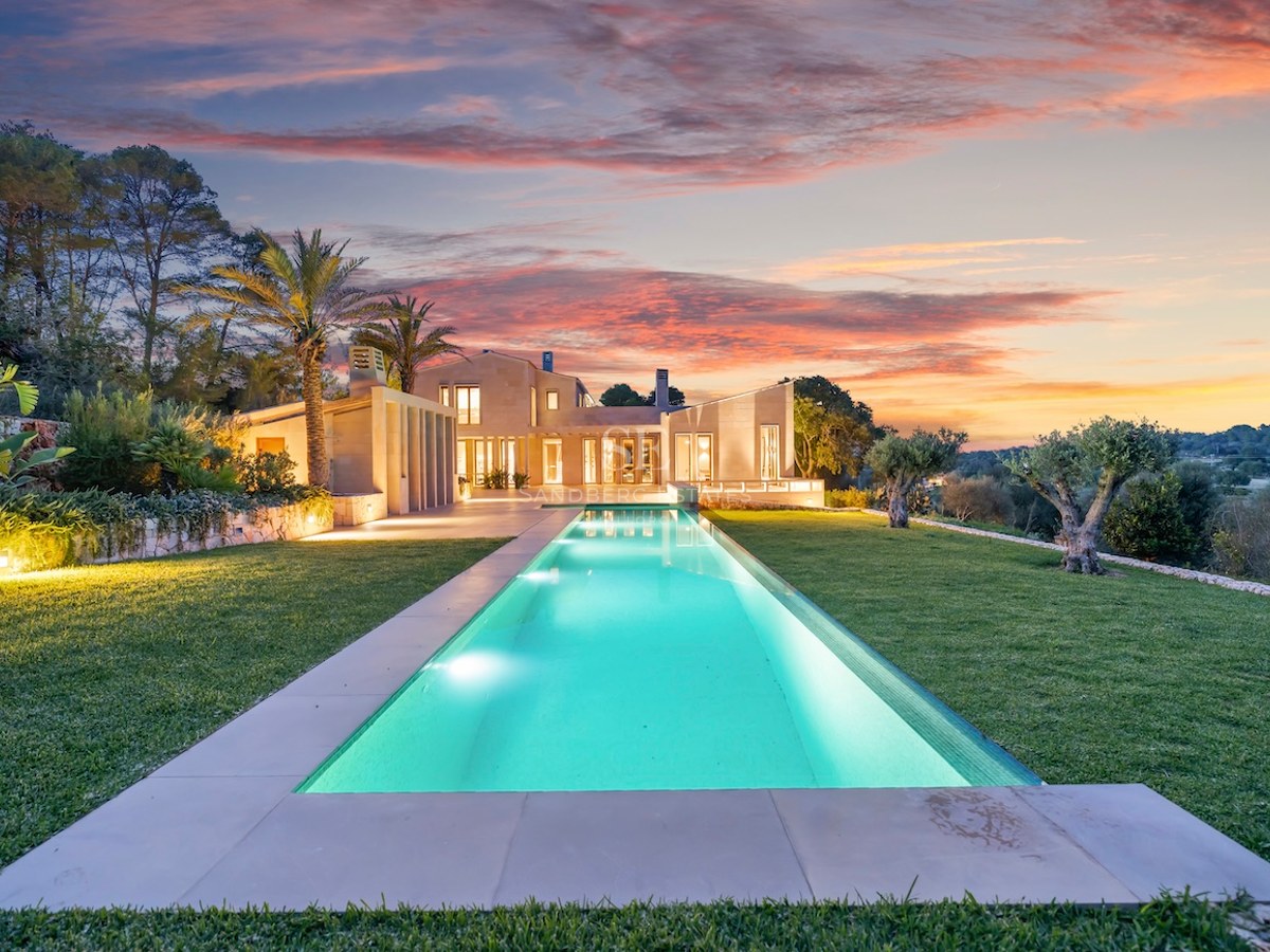 A long, illuminated rectangular pool leading to a modern stone villa under a vibrant pink and orange sunset sky.