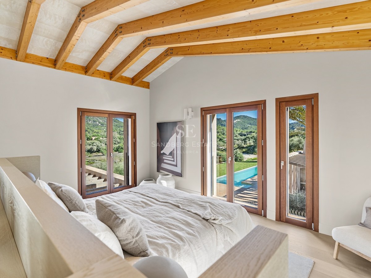 Modern bedroom with vaulted wood beam ceiling, bed with beige linens, and glass doors overlooking a pool and green hills.