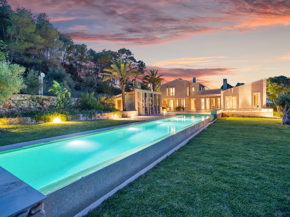 Long glowing lap pool leading to a modern stone villa under a vibrant orange and pink sunset sky.