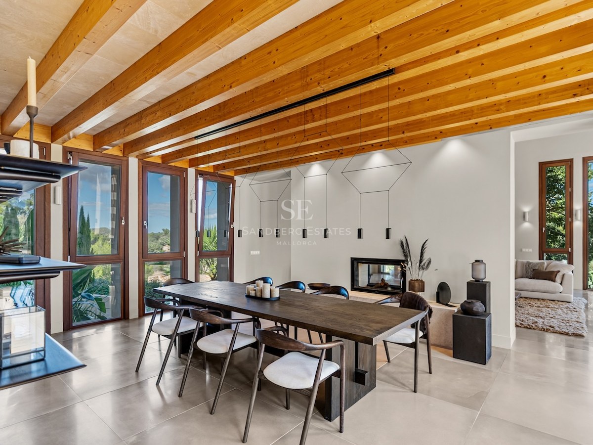 Large wooden dining table under a beamed ceiling with floor-to-ceiling windows showing nature views.