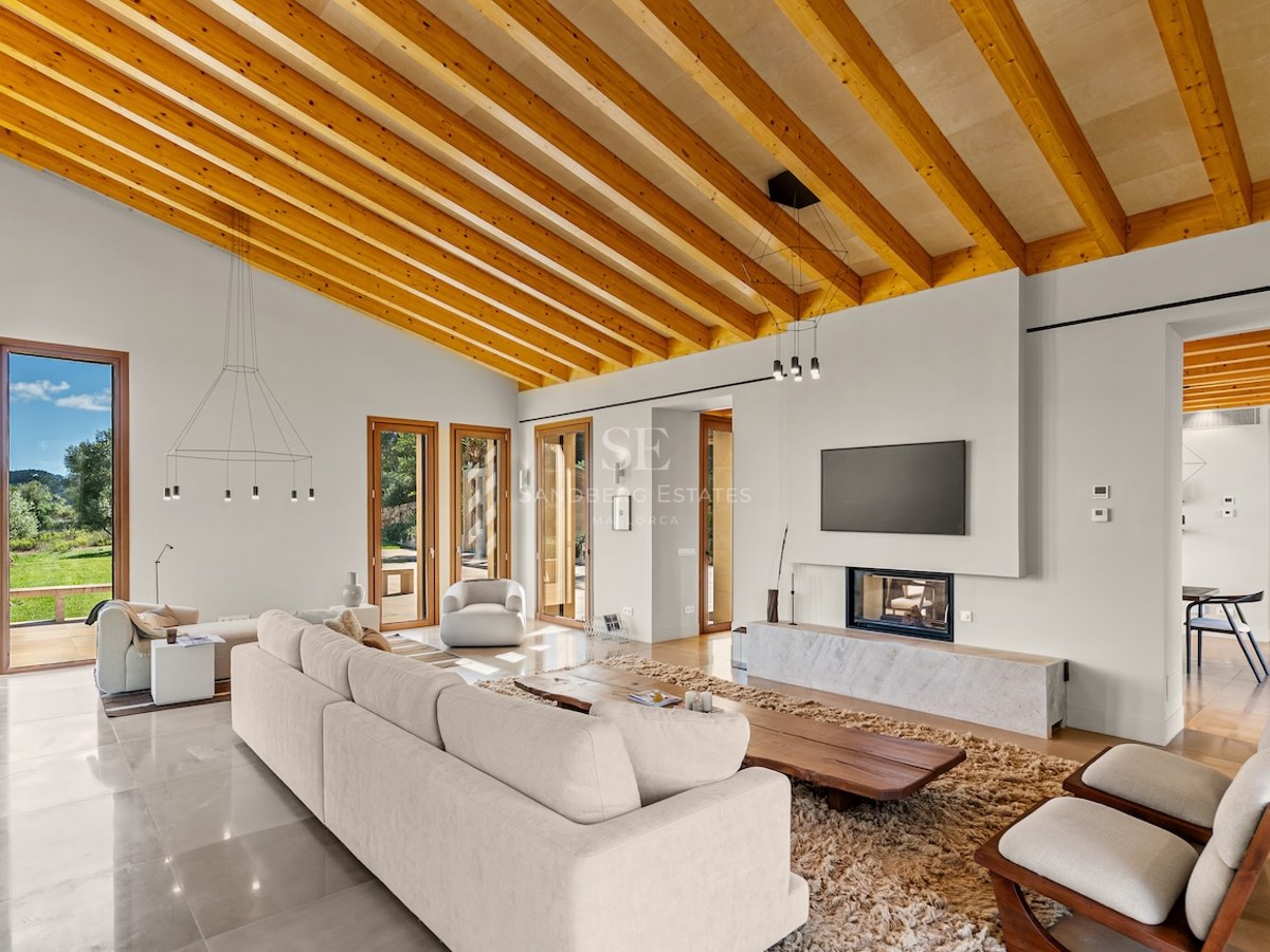 Spacious modern living room featuring high ceilings with wood beams, a large sofa, fireplace, and garden views.
