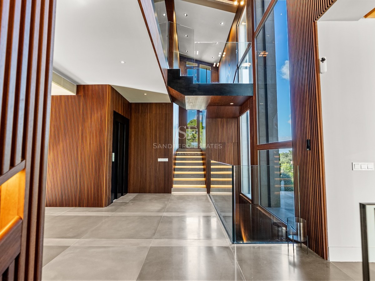Modern foyer featuring slatted wood walls, a glass-railed staircase with LED lighting, and massive floor-to-ceiling windows.