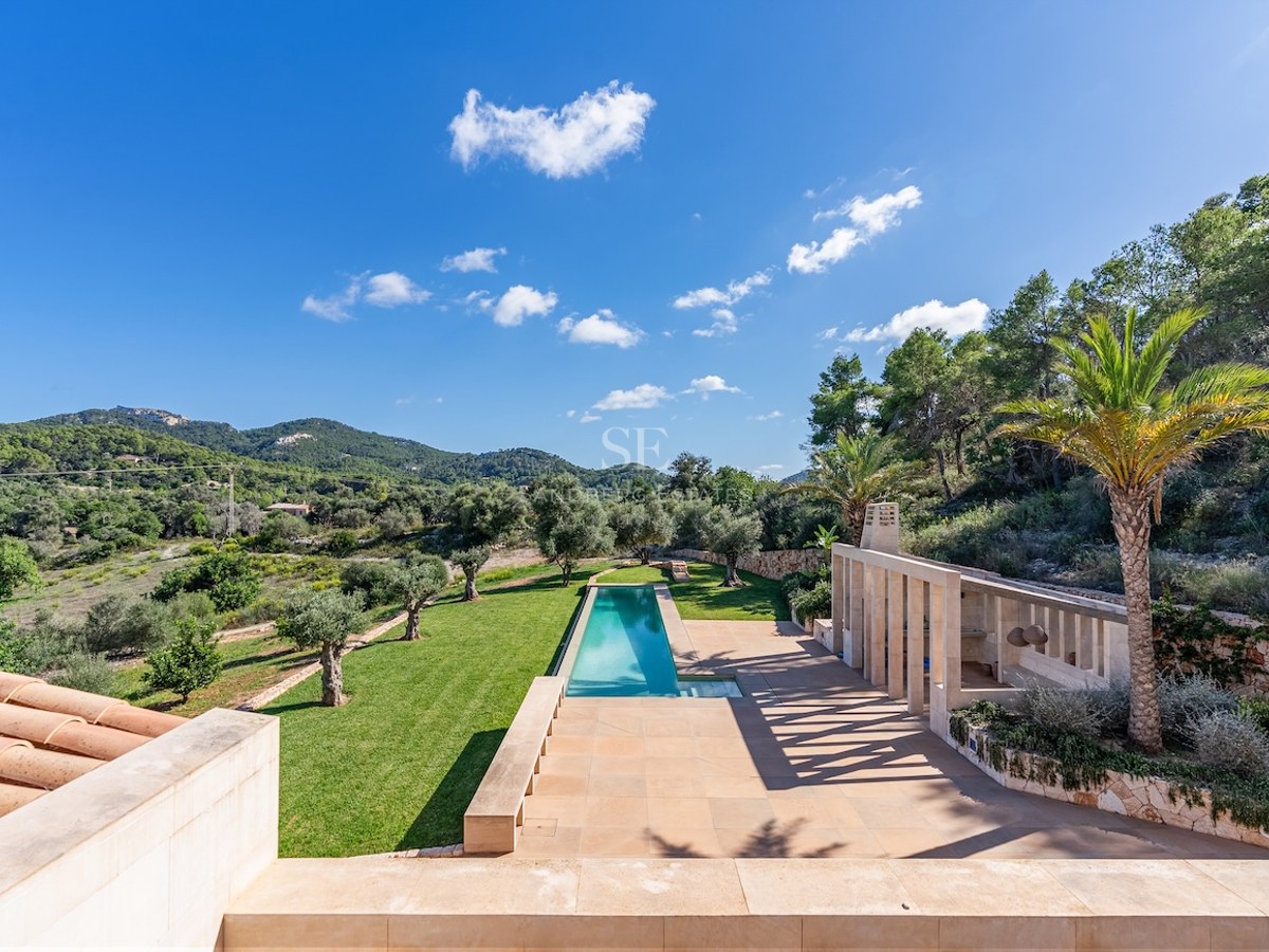 High-angle view of a long turquoise lap pool, stone terrace, and olive trees set against lush green hills and a blue sky.
