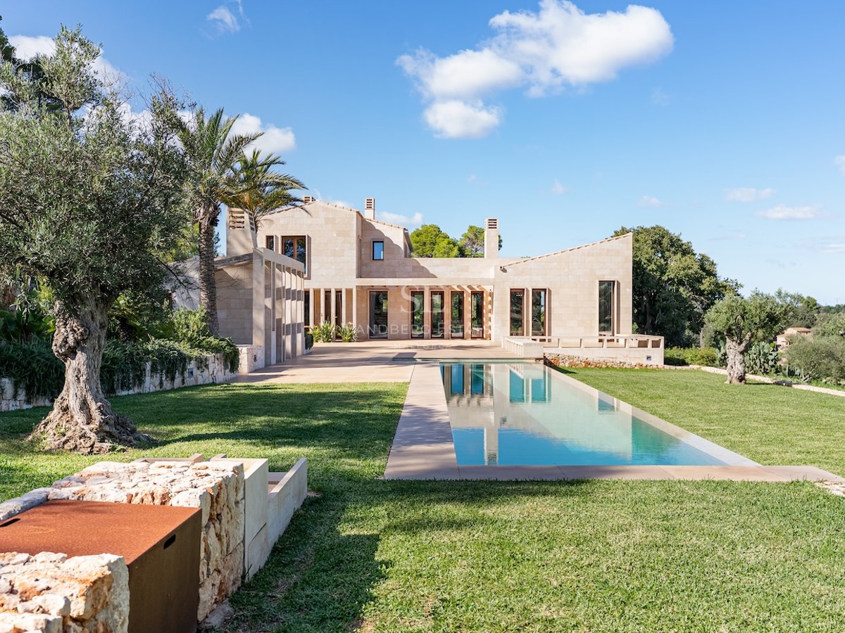 Modern sandstone villa with a long rectangular lap pool, lush green lawn, and ancient olive trees under a blue sky.