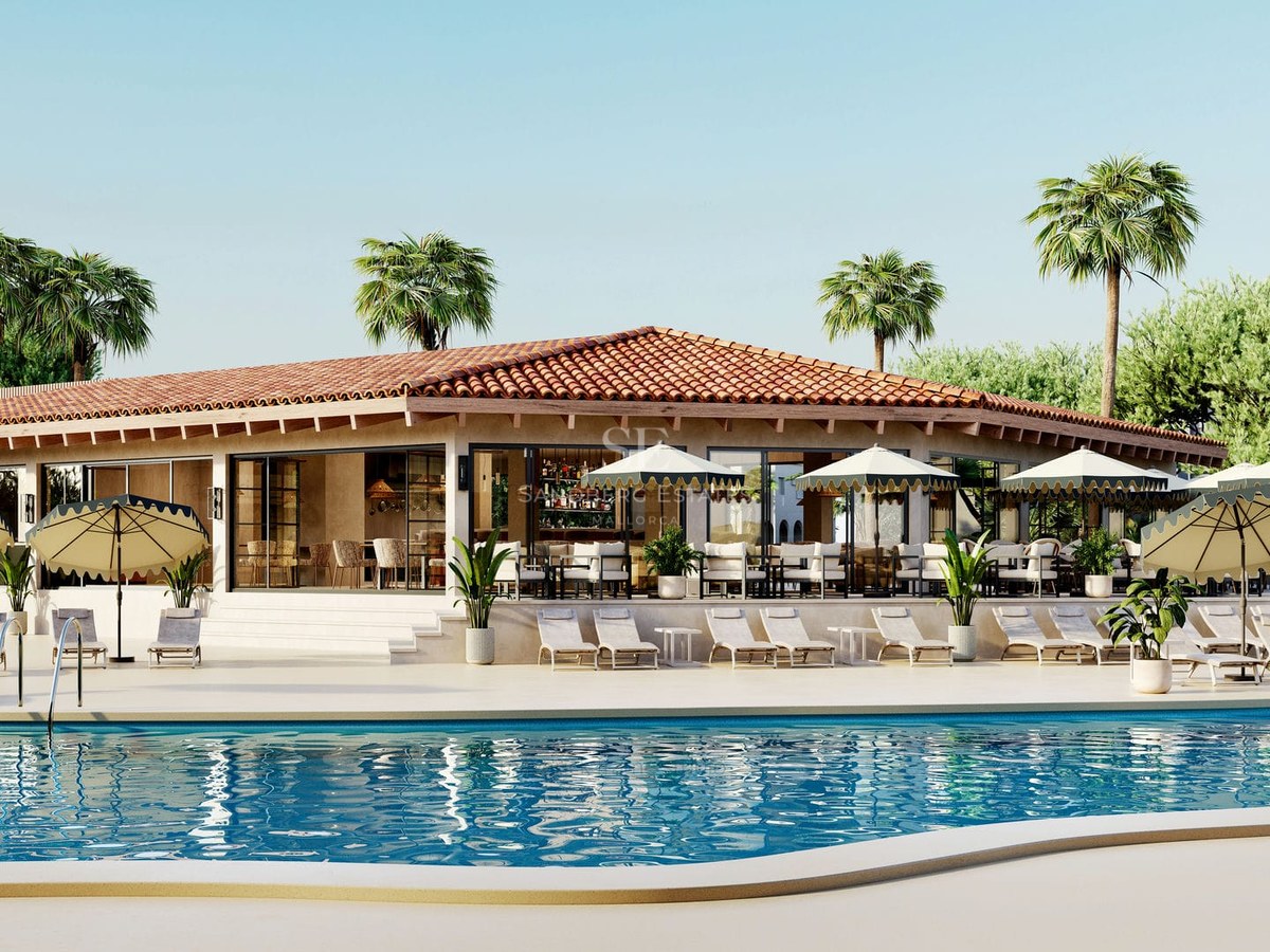Large blue swimming pool with sun loungers and umbrellas in front of a Mediterranean-style building with palms.