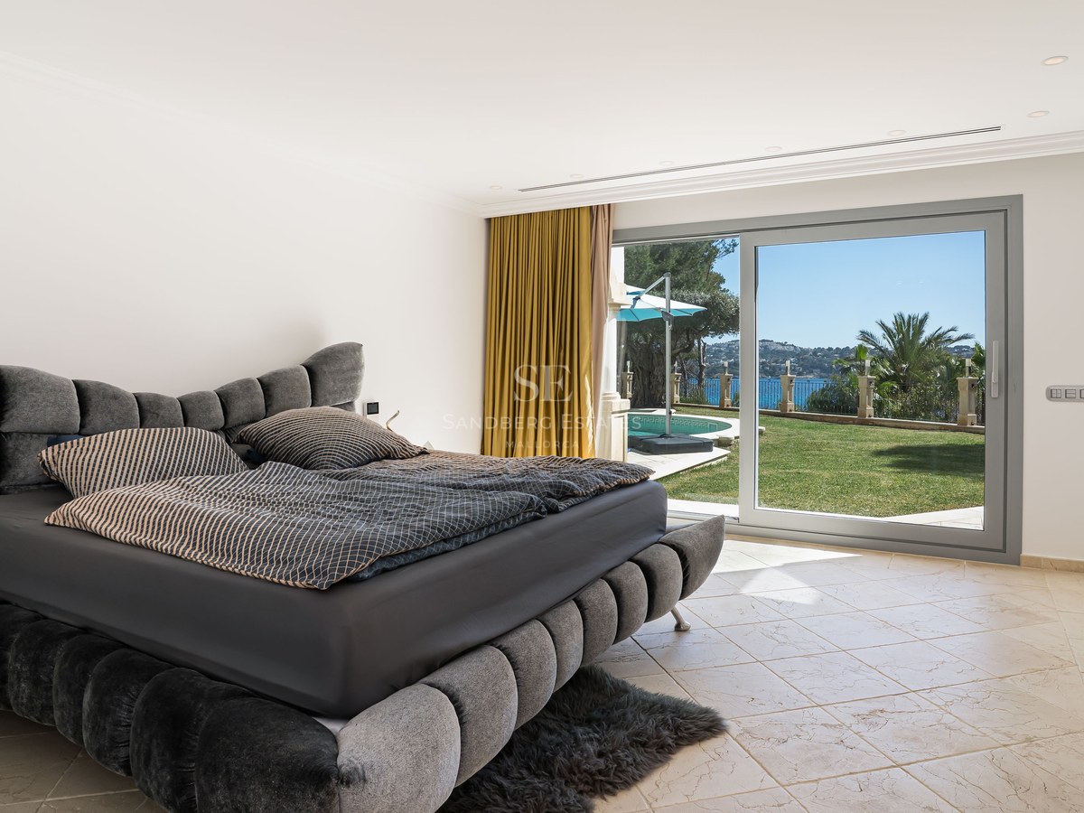 Chambre moderne avec lit gris capitonné et grandes baies vitrées ouvrant sur un jardin, une piscine et la mer.
