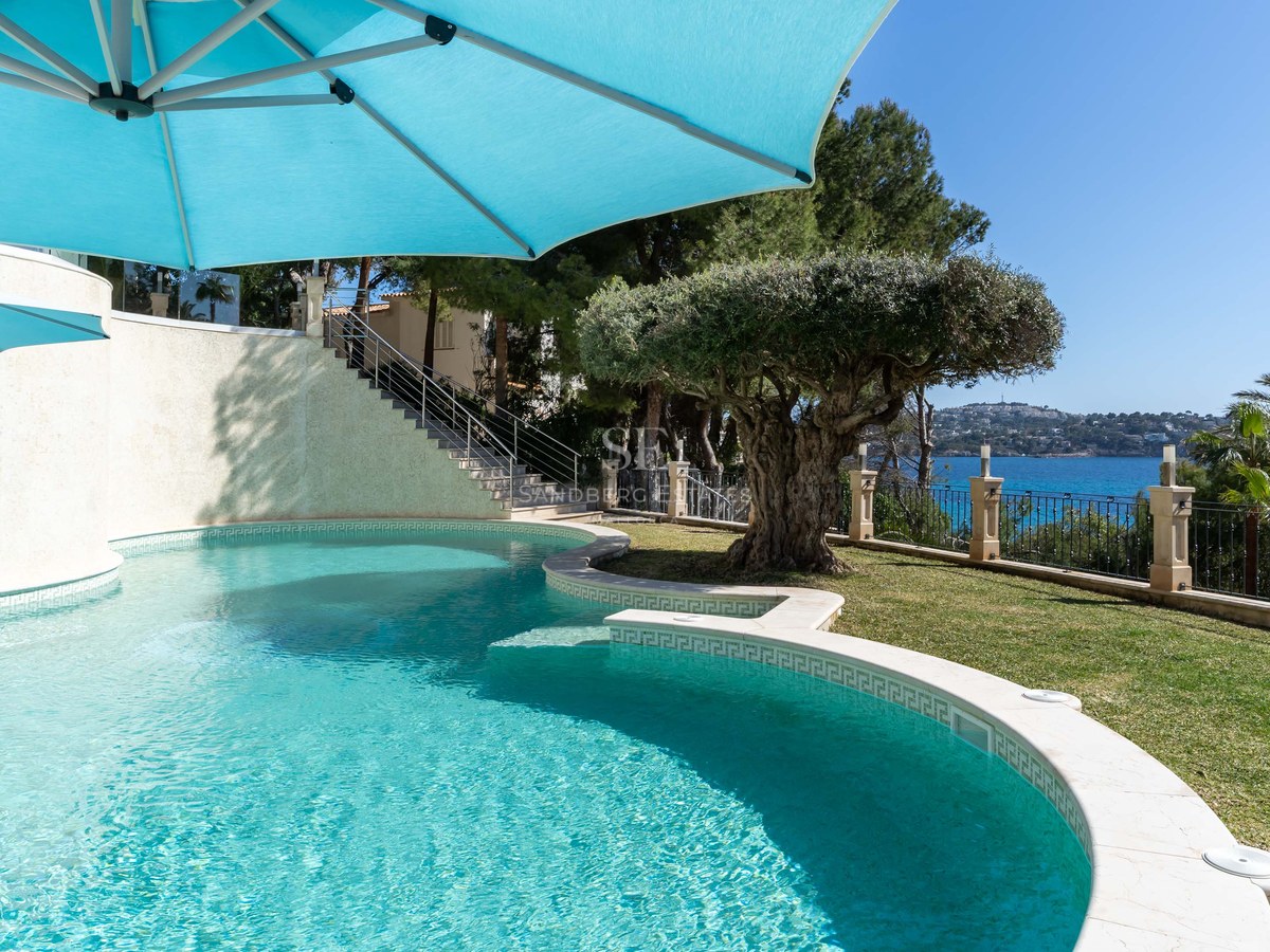 Piscine courbée aux eaux turquoise avec un olivier et vue sur la mer sous un parasol bleu.
