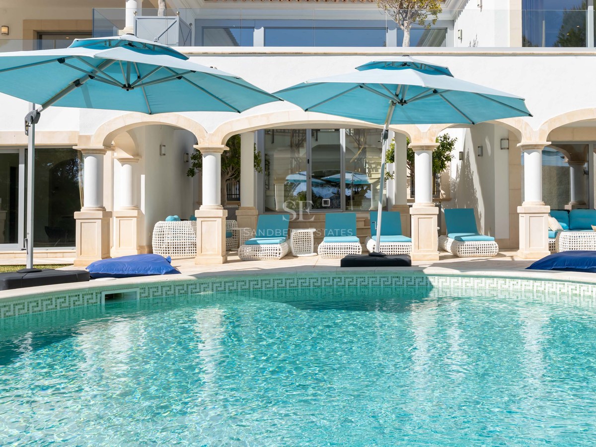 Piscine turquoise avec colonnes blanches classiques et parasols bleu canard sur une terrasse ensoleillée.