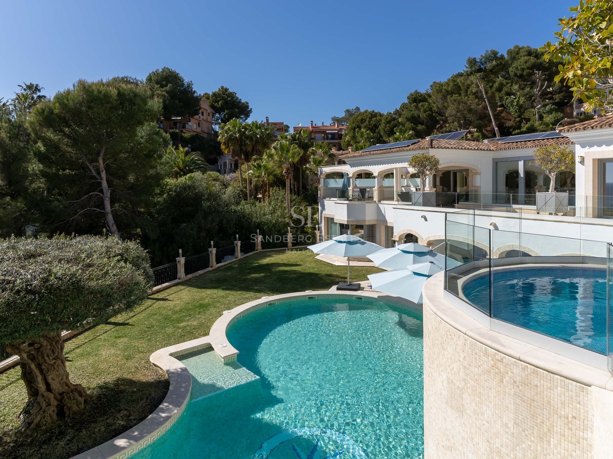 Villa de luxe avec piscine turquoise incurvée, garde-corps en verre et végétation dense à Majorque.