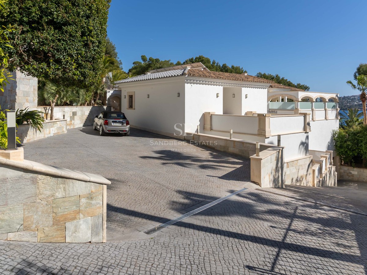 Villa méditerranéenne blanche avec allée pavée privée, murs en pierre et voiture garée sous un ciel bleu.