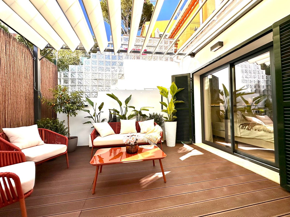 Stylish outdoor terrace with red and white furniture, orange table, lush plants, and a white retractable awning in the sun.