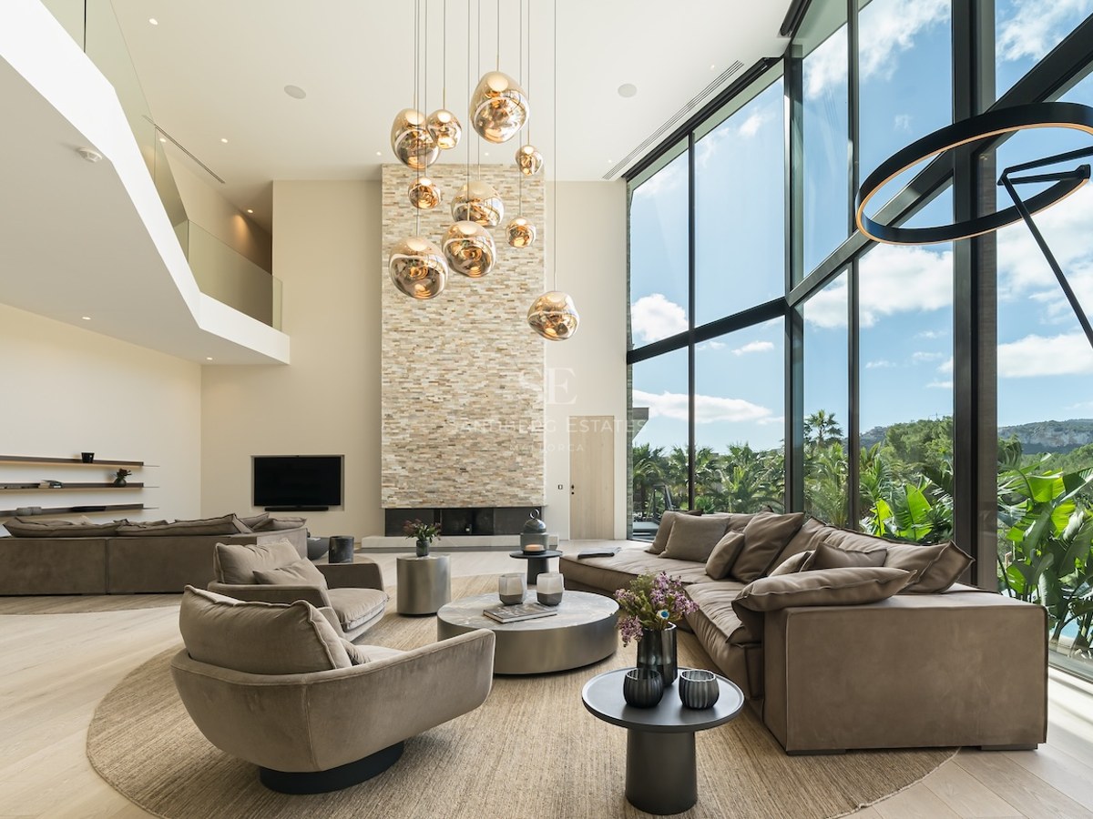 Un salon moderne à double hauteur avec cheminée en pierre, suspensions dorées et grandes baies vitrées sur le jardin.