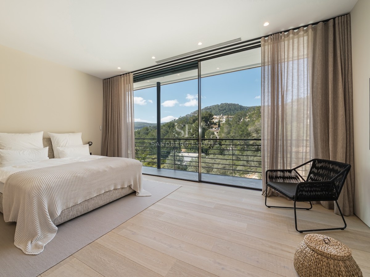 Dormitorio moderno con suelos de madera clara, grandes ventanales de suelo a techo y vistas a colinas verdes.