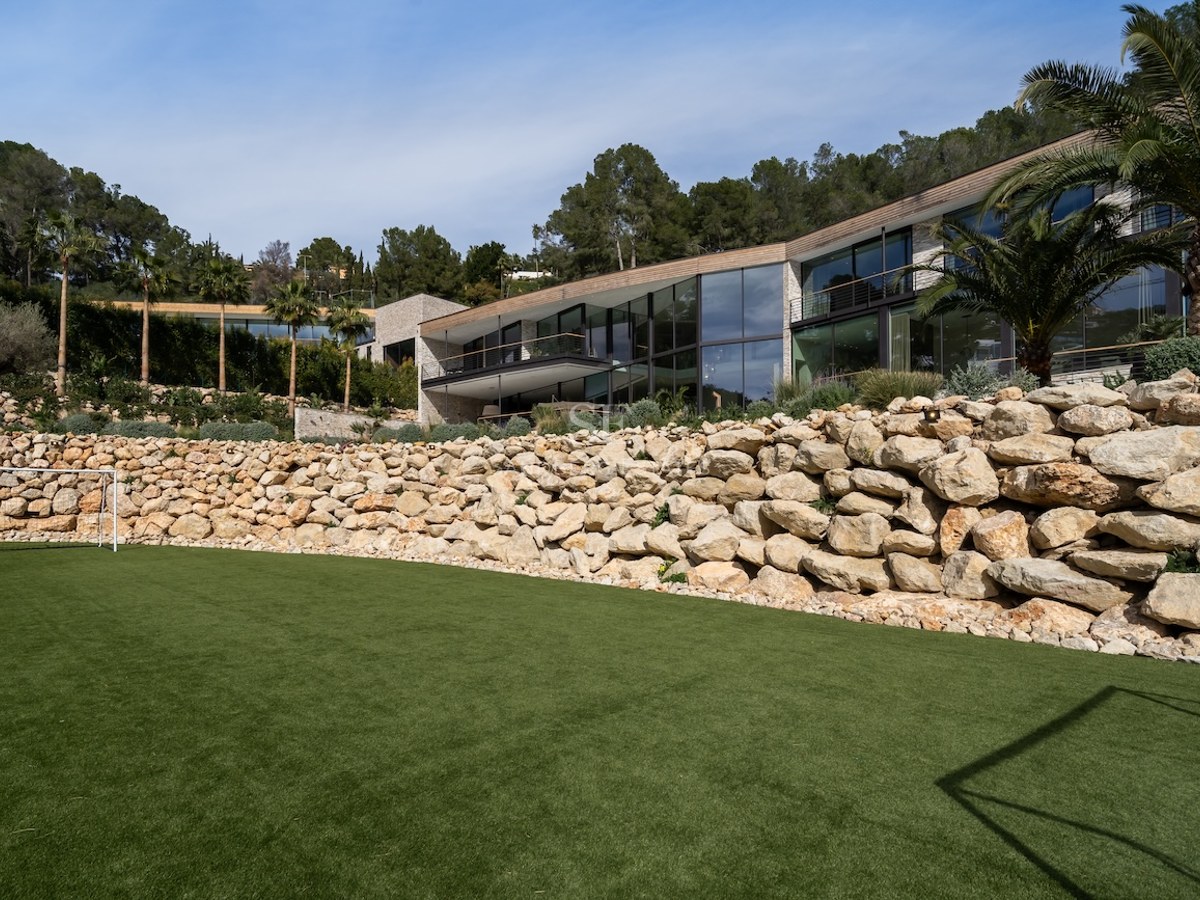 Façade de villa contemporaine avec baies vitrées derrière un mur de soutènement en pierre naturelle et pelouse verte.
