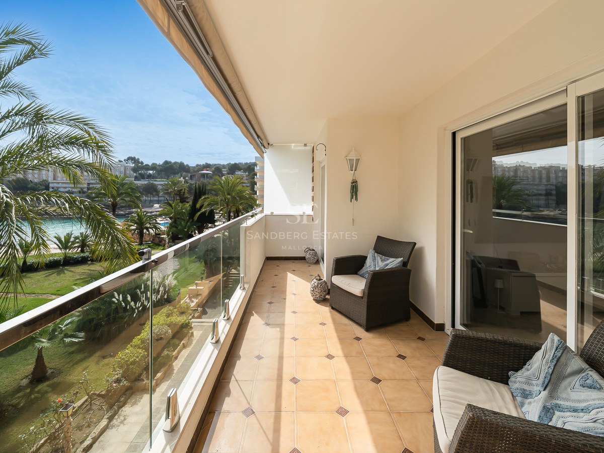 Balcon ensoleillé avec chaises en osier, garde-corps en verre et vue sur des palmiers et jardins.