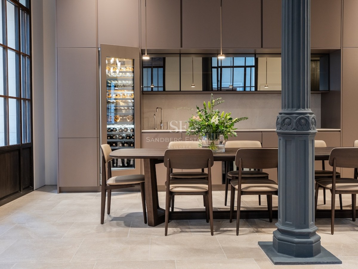 Comedor moderno con mesa de madera, sillas beige, columna industrial azul y vinoteca de cristal integrada.