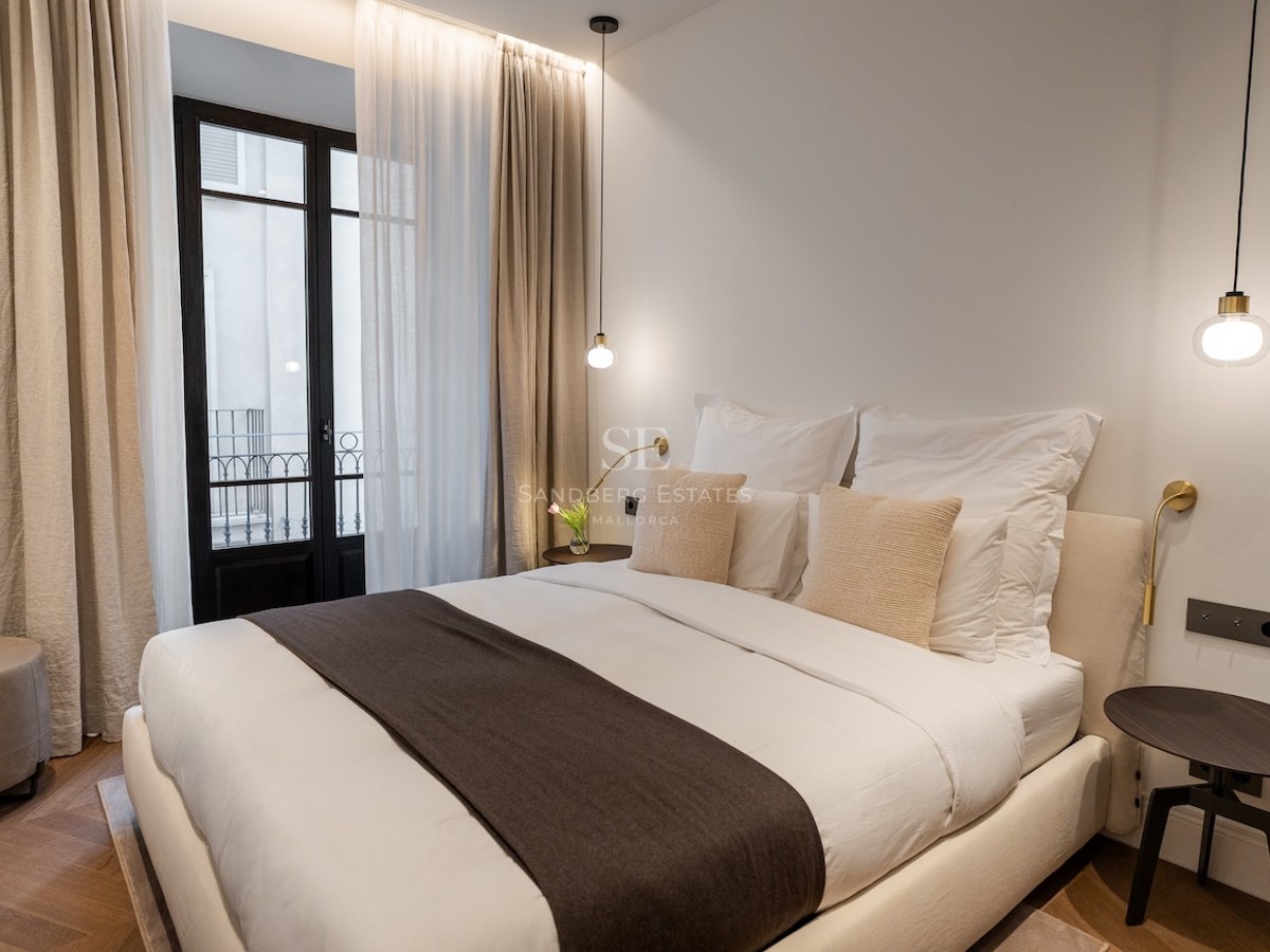 Dormitorio luminoso con cama doble, suelos de madera en espiga, lámparas colgantes y balcón.