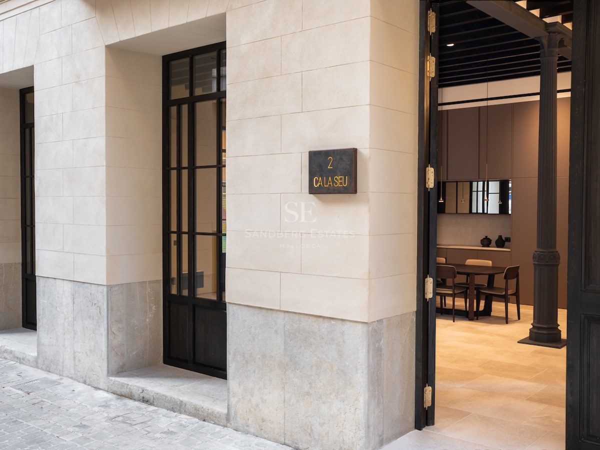 Primer plano de la entrada de un edificio moderno con bloques de piedra crema, puerta de metal negro y placa C'an Seu 2.