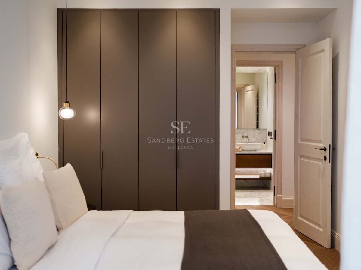 Dormitorio moderno con armario taupe, ropa de cama blanca y vista a un baño de mármol a través de una puerta abierta.